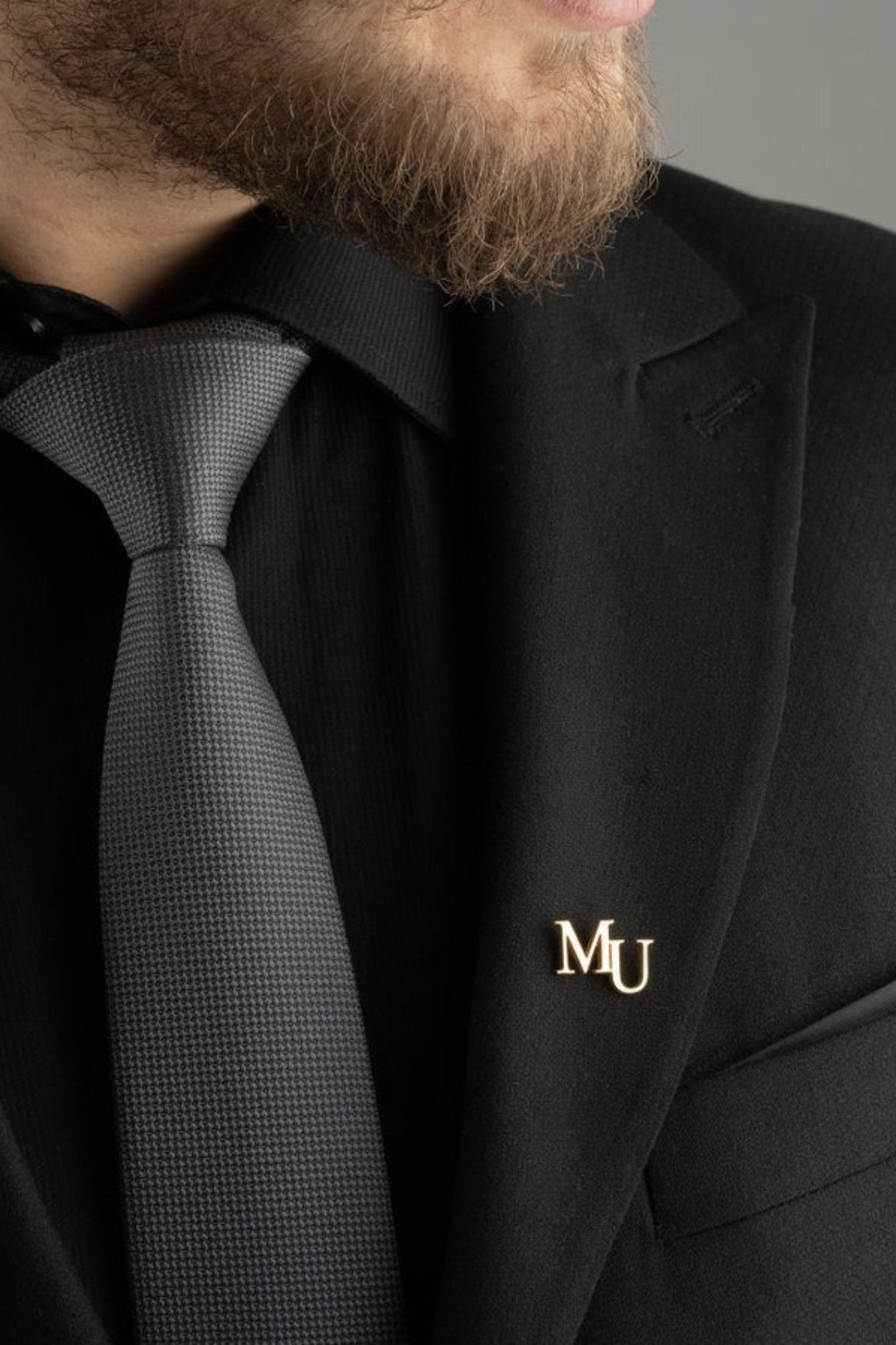 Man wearing a black suit with a gray tie and a gold 'MU' lapel pin on a gray background